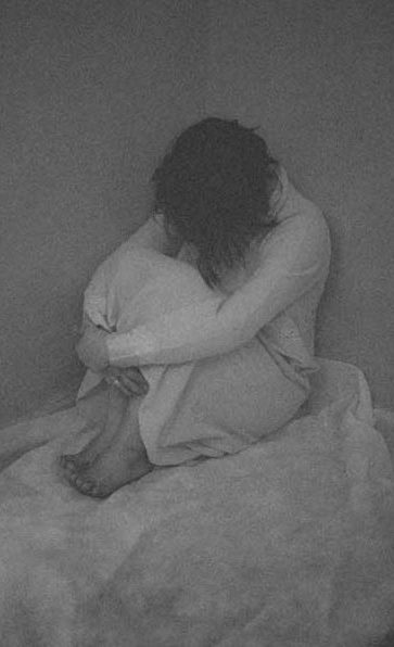 This woman is sitting on a bed in a corner, with her head down, holding her knees. Women are often taught to act smaller. Judgement placed on us is multifaceted.